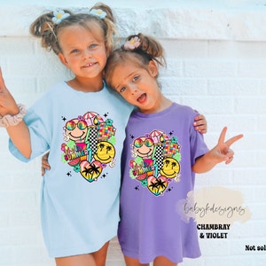 May include: Two children wearing light blue and purple t-shirts. Each shirt features a vibrant graphic with smiley faces, sunglasses, a disco ball, and the word "SUMMER". The t-shirts have a loose, comfortable fit.