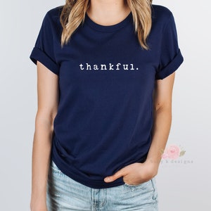 Op de afbeelding: Een marineblauw t-shirt met het woord "thankful" in het wit gedrukt. Het shirt heeft opgerolde mouwen en een klassieke ronde hals. Het model draagt een blauwe spijkerbroek.