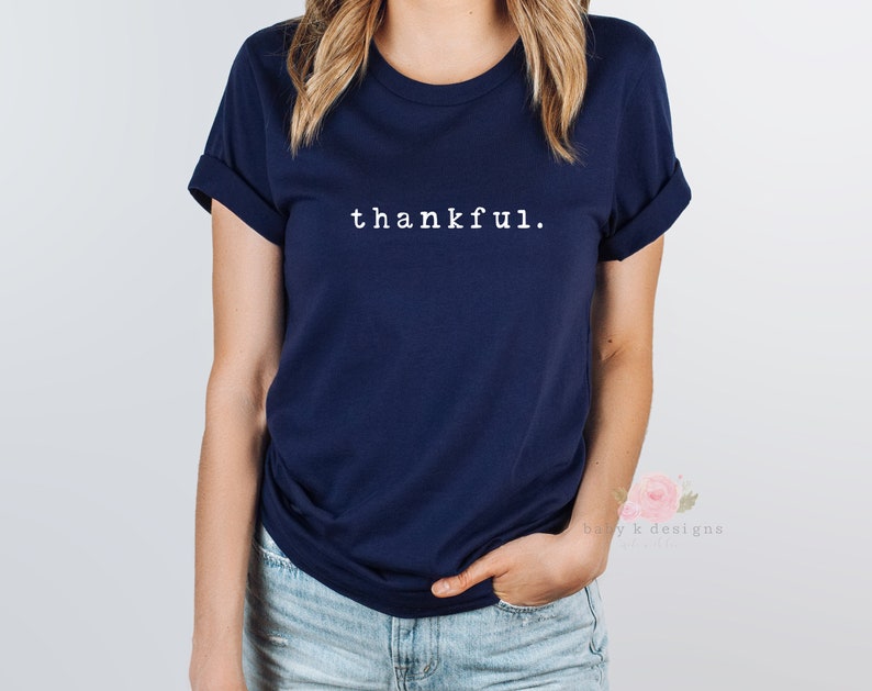 Op de afbeelding: Een marineblauw t-shirt met het woord "thankful" in het wit gedrukt. Het shirt heeft opgerolde mouwen en een klassieke ronde hals. Het model draagt een blauwe spijkerbroek.