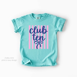 May include: A chalky mint short-sleeve t-shirt featuring a graphic design with vertical light pink stripes and the navy blue text "club ten" written in a cursive script, positioned above a navy blue bow illustration on the front center.