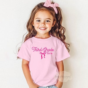 May include: A light pink t-shirt with the words "Third Grade" and the name "Sofia" in a cursive font, along with a bow graphic. The girl in the image is wearing a pink bow in her hair.