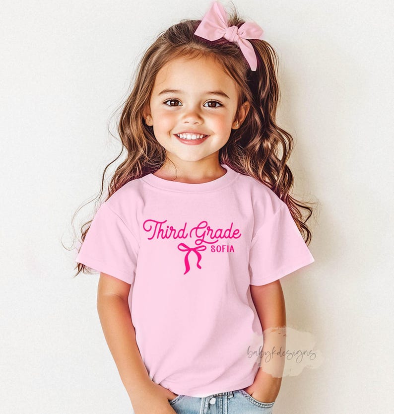 May include: A light pink t-shirt with the words "Third Grade" and the name "Sofia" in a cursive font, along with a bow graphic. The girl in the image is wearing a pink bow in her hair.
