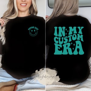 May include: Black sweatshirt with a teal smiley face with stars and a lightning bolt on the left chest and the text "IN MY CUSTOM ERA" in teal retro font on the back.