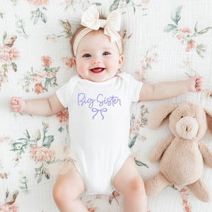 May include: A baby wearing a white onesie with "Big Sister" in lavender script and a bow graphic. The baby is lying on a floral patterned blanket next to a plush bunny toy. A cream-colored bow headband completes the outfit.