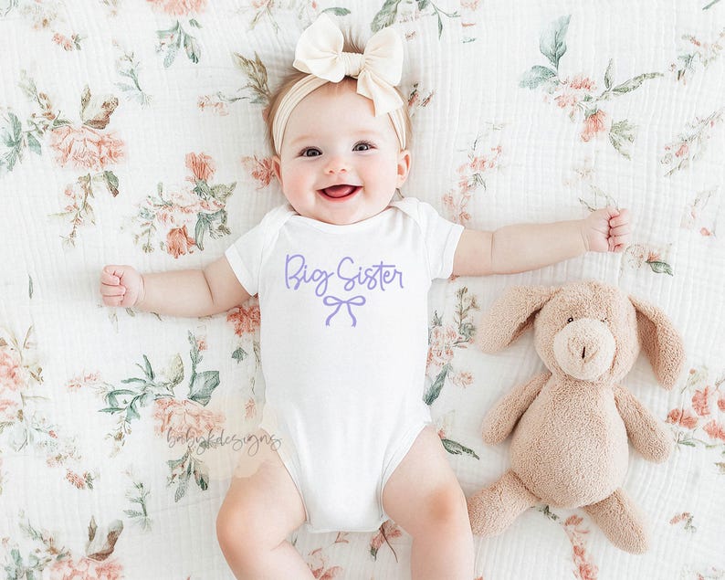 May include: A baby wearing a white onesie with "Big Sister" in lavender script and a bow graphic. The baby is lying on a floral patterned blanket next to a plush bunny toy. A cream-colored bow headband completes the outfit.