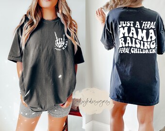 May include: Black oversized t-shirt with a white graphic of a skeleton hand making a rock and roll sign with the text "Feral Mama" on the front. The back of the shirt has white text that reads "Just a Feral Mama Raising Feral Children".