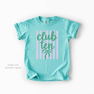 May include: A chalky mint short-sleeve t-shirt featuring a central graphic of vertical lavender stripes. Overlaid on the stripes is the text "club ten" in a dark green cursive font, positioned above a small, decorative green bow illustration.
