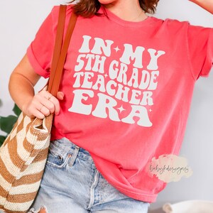 May include: A pink t-shirt with the text "In My 5th Grade Teacher Era" in white retro-style lettering with stars.