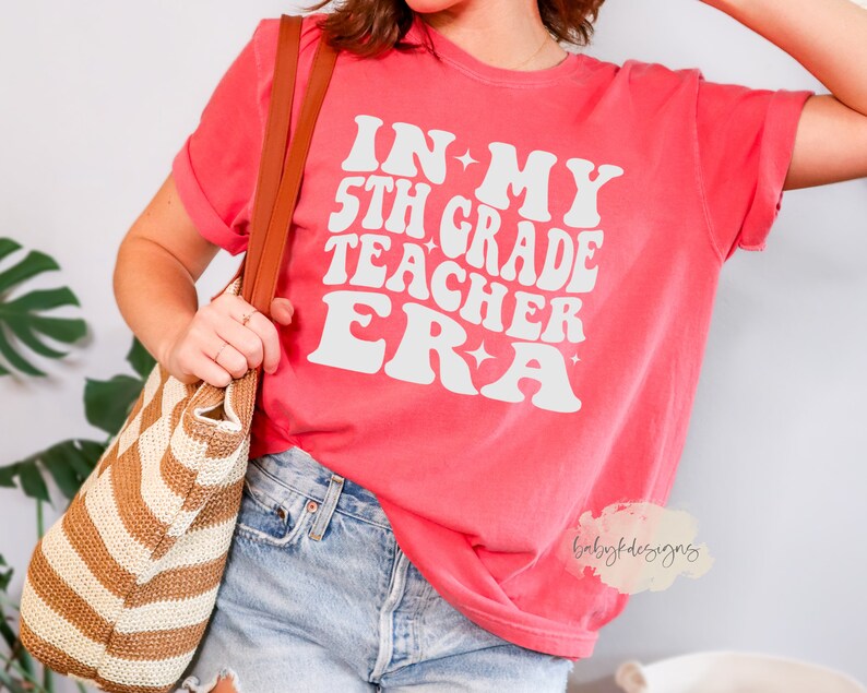 May include: A pink t-shirt with the text "In My 5th Grade Teacher Era" in white retro-style lettering with stars.