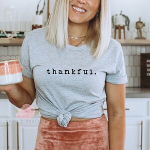 Op de afbeelding: Lichtgrijs t-shirt met het woord "thankful" in zwart schrift. Het shirt is in de taille geknoopt. Een mok met een wit en koraal ontwerp wordt in de hand gehouden. Het thema is dankbaarheid en vrijetijdskleding.
