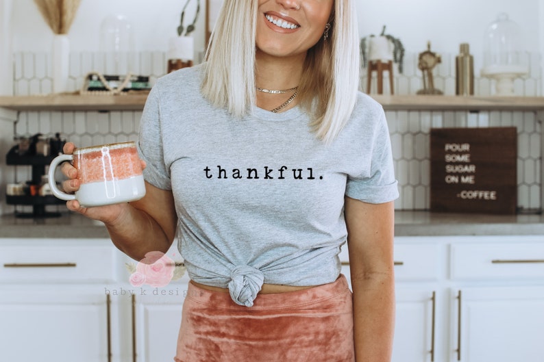 Op de afbeelding: Lichtgrijs t-shirt met het woord "thankful" in zwart schrift. Het shirt is in de taille geknoopt. Een mok met een wit en koraal ontwerp wordt in de hand gehouden. Het thema is dankbaarheid en vrijetijdskleding.