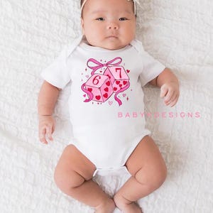 May include: A white baby onesie with a pink and red heart-themed dice graphic. The dice show the numbers six and seven. The text "BABY DESIGNS" is printed in pink on the onesie.