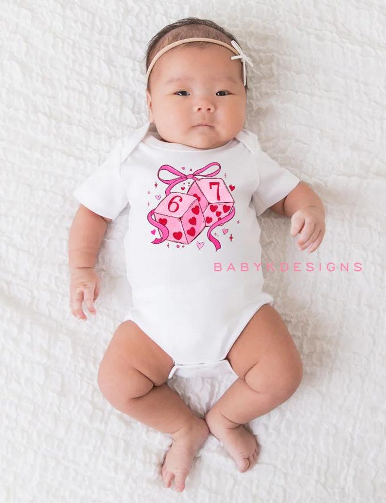 May include: A white baby onesie with a pink and red heart-themed dice graphic. The dice show the numbers six and seven. The text "BABY DESIGNS" is printed in pink on the onesie.