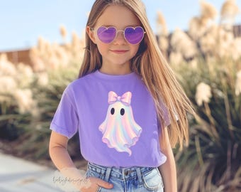 Puede incluir: Una camiseta de color lila claro con un diseño de fantasma y un lazo. El fantasma tiene un esquema de color arcoíris pastel. La camiseta se combina con vaqueros azules y gafas de sol en forma de corazón.