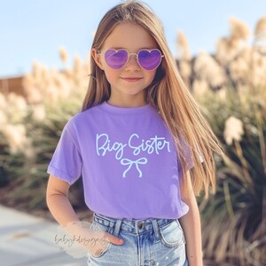May include: A young person wearing heart-shaped sunglasses and a purple t-shirt with the words "Big Sister" in light blue script. The shirt also features a bow graphic. The person is wearing blue denim shorts.