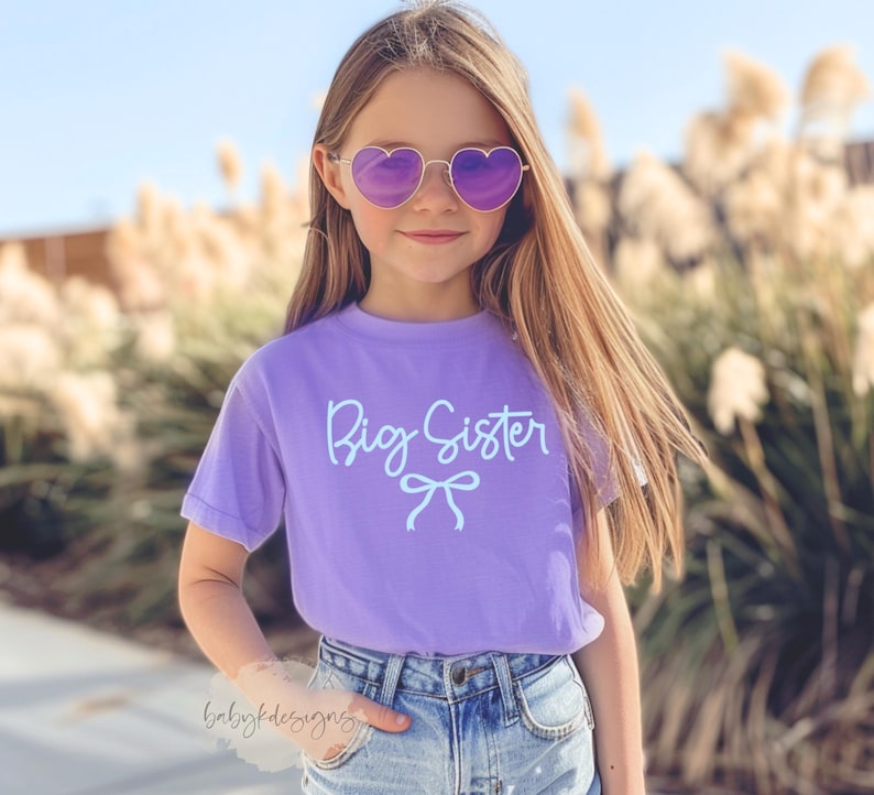 May include: A young person wearing heart-shaped sunglasses and a purple t-shirt with the words "Big Sister" in light blue script. The shirt also features a bow graphic. The person is wearing blue denim shorts.