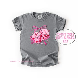 May include: A gray t-shirt with a graphic of two pink dice, one showing a 6 and the other a 7, decorated with hearts and a pink ribbon. The text "Comfort Colors Youth & Adults Sizes" is in a pink circle.