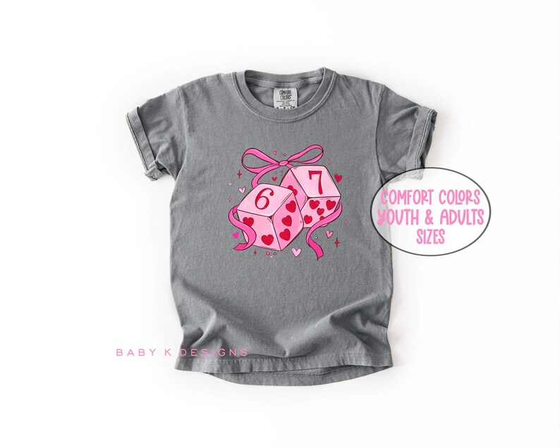 May include: A gray t-shirt with a graphic of two pink dice, one showing a 6 and the other a 7, decorated with hearts and a pink ribbon. The text "Comfort Colors Youth & Adults Sizes" is in a pink circle.