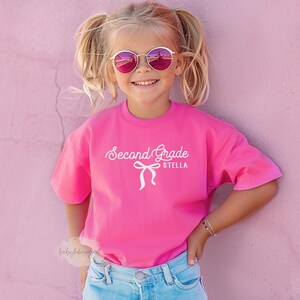 May include: A young girl wearing a bright pink t-shirt with the words "Second Grade Stella" in white script. She is also wearing round, pink-tinted sunglasses and blue jeans. The background is a solid pink wall.