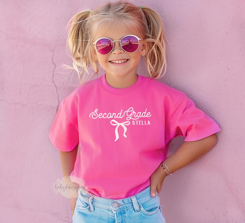 May include: A young girl wearing a bright pink t-shirt with the words "Second Grade Stella" in white script. She is also wearing round, pink-tinted sunglasses and blue jeans. The background is a solid pink wall.