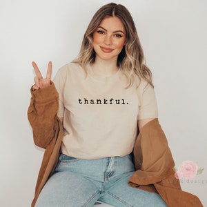 Op de afbeelding: Een beige t-shirt met het woord "thankful" in het zwart gedrukt. De persoon draagt een blauwe spijkerbroek en een bruine jas. De persoon steekt twee vingers op als vredesteken.