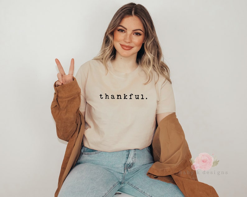 Op de afbeelding: Een beige t-shirt met het woord "thankful" in het zwart gedrukt. De persoon draagt een blauwe spijkerbroek en een bruine jas. De persoon steekt twee vingers op als vredesteken.