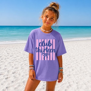 May include: A lavender short-sleeved t-shirt featuring a graphic design with vertical light pink stripes. The text "club thirteen" is printed in dark blue cursive script above a dark blue bow illustration centered on the front of the shirt.