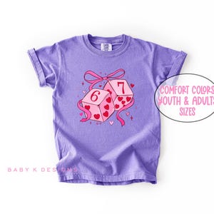 May include: A lilac-colored t-shirt featuring a graphic of two dice with the numbers 6 and 7, decorated with hearts and a pink ribbon. The text "Comfort Colors Youth & Adults Sizes" is in a white oval. The brand name "Baby K Designs" is at the bottom.