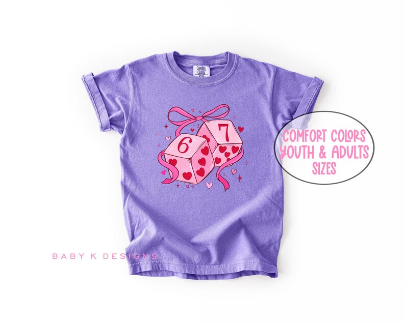 May include: A lilac-colored t-shirt featuring a graphic of two dice with the numbers 6 and 7, decorated with hearts and a pink ribbon. The text "Comfort Colors Youth & Adults Sizes" is in a white oval. The brand name "Baby K Designs" is at the bottom.