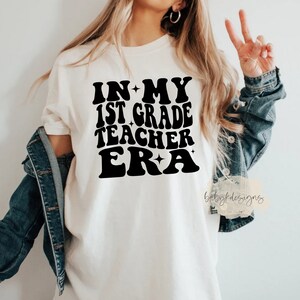 May include: White t-shirt with black text that reads "In My 1st Grade Teacher Era" in a retro font with stars.