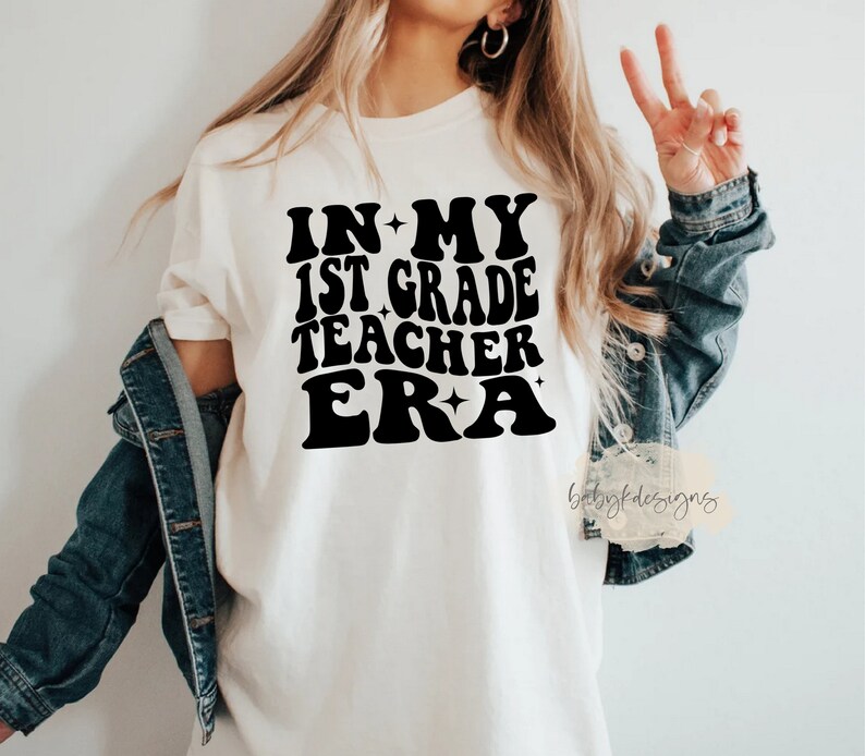 May include: White t-shirt with black text that reads "In My 1st Grade Teacher Era" in a retro font with stars.