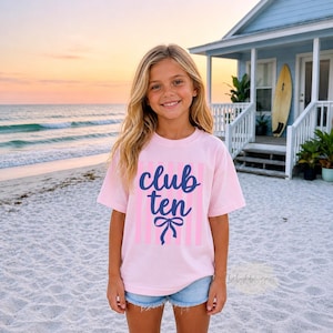 May include: A light pink short-sleeved t-shirt featuring vertical light pink stripes and the dark blue text "club ten" written in a cursive script, positioned above a dark blue graphic of a bow tied in a ribbon style.