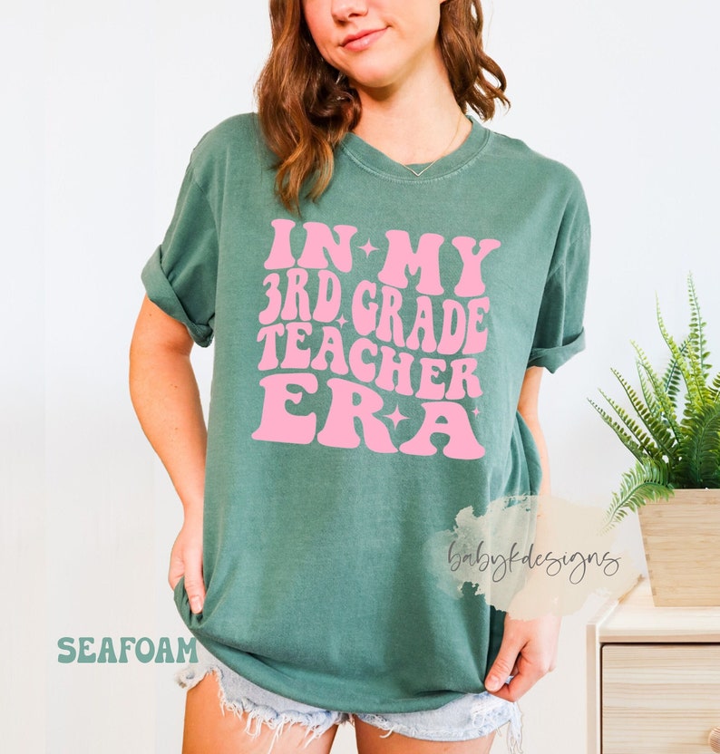 May include: A teal blue t-shirt with the text "IN MY 3RD GRADE TEACHER ERA" in pink lettering with stars. The text is in a retro font style.