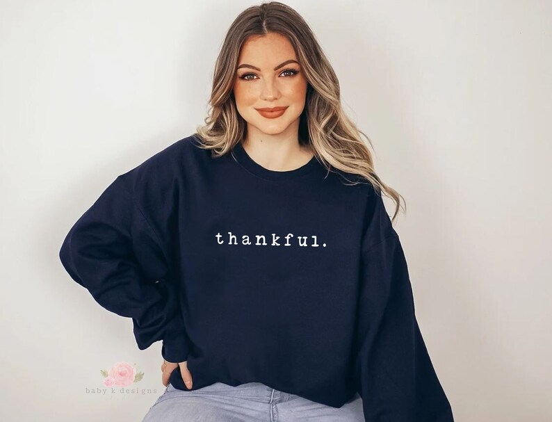 Op de afbeelding: Marineblauwe sweatshirt met het woord "thankful" in het wit in het midden. De tekst is in een eenvoudige lettertype. De sweatshirt heeft lange mouwen en een ronde hals.