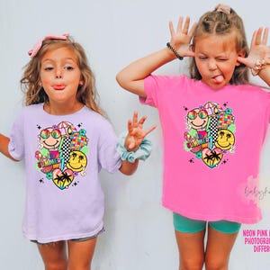 May include: Two young girls wearing matching t-shirts with a colourful summer-themed graphic. The shirts are light purple and bright pink, featuring smiley faces, sunglasses, and the words "SUMMER". The girls are making silly faces.
