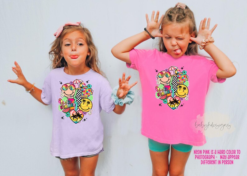 May include: Two young girls wearing matching t-shirts with a colourful summer-themed graphic. The shirts are light purple and bright pink, featuring smiley faces, sunglasses, and the words "SUMMER". The girls are making silly faces.
