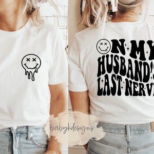 May include: White t-shirt with a dripping smiley face on the front and the text "On my husband's last nerve" on the back.