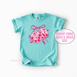 May include: A turquoise t-shirt featuring a graphic of two pink dice with the numbers 6 and 7, adorned with hearts and a pink ribbon. The shirt has the text "Comfort Colors Youth & Adults Sizes".