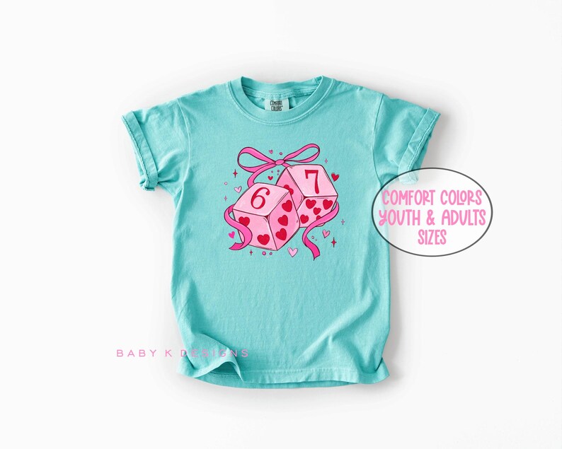May include: A turquoise t-shirt featuring a graphic of two pink dice with the numbers 6 and 7, adorned with hearts and a pink ribbon. The shirt has the text "Comfort Colors Youth & Adults Sizes".