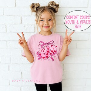 May include: A pink t-shirt featuring a graphic of two dice with the numbers 6 and 7, decorated with hearts and a pink ribbon. The text "COMFORT COLORS YOUTH & ADULTS SIZES" is in a circle. The shirt is worn by a young girl.