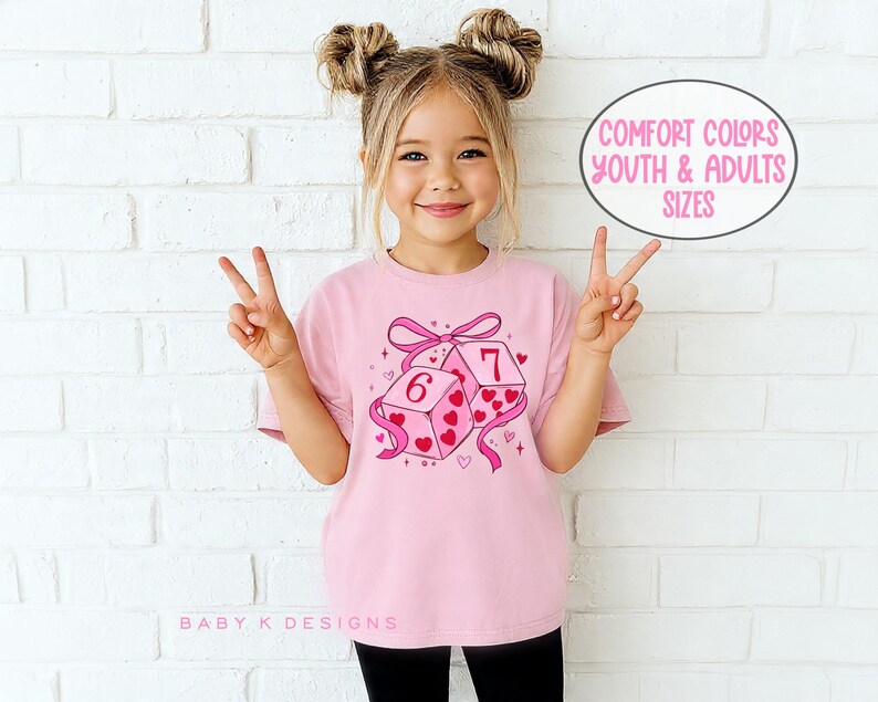 May include: A pink t-shirt featuring a graphic of two dice with the numbers 6 and 7, decorated with hearts and a pink ribbon. The text "COMFORT COLORS YOUTH & ADULTS SIZES" is in a circle. The shirt is worn by a young girl.