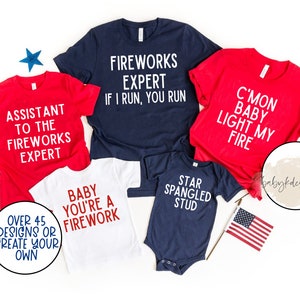 Op de afbeelding: Vijf rood, wit en blauwe shirts met tekst die luidt "Assistant to the Fireworks Expert", "Fireworks Expert If I Run, You Run", "C'mon Baby Light My Fire", "Baby You're a Firework", en "Star Spangled Stud".