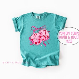 May include: Teal-colored t-shirt featuring a graphic of two dice with the numbers 6 and 7, decorated with pink hearts and a pink ribbon. The text "Comfort Colors Youth & Adults Sizes" is in a white oval. The shirt has rolled-up sleeves.