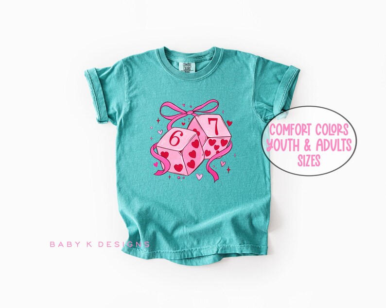 May include: Teal-colored t-shirt featuring a graphic of two dice with the numbers 6 and 7, decorated with pink hearts and a pink ribbon. The text "Comfort Colors Youth & Adults Sizes" is in a white oval. The shirt has rolled-up sleeves.