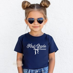 May include: A young child wearing a navy blue t-shirt with the words "First Grade Hannah" in white script. The child is also wearing heart-shaped sunglasses with a gold frame.
