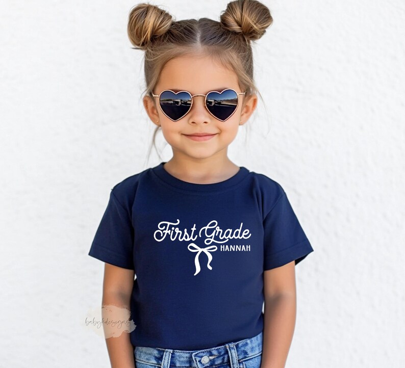 May include: A young child wearing a navy blue t-shirt with the words "First Grade Hannah" in white script. The child is also wearing heart-shaped sunglasses with a gold frame.