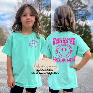 May include: A turquoise blue t-shirt with a pink smiley face and the text "I hope you have a good day" on the front. The back of the shirt has the text "Dear person behind me I hope you have a good day Lots of love the person in front of you" in pink.