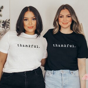 Op de afbeelding: Twee mensen dragen t-shirts met het woord "thankful." op de voorkant. E&eacute;n shirt is wit, de andere is zwart. De tekst is in een eenvoudig, schreefloos lettertype. De mensen staan voor een neutrale achtergrond.