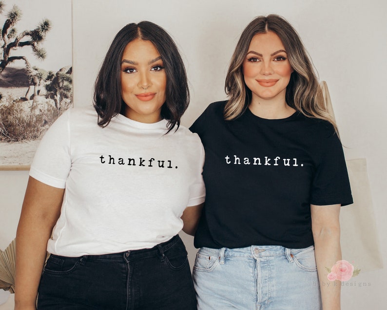 Op de afbeelding: Twee mensen dragen t-shirts met het woord "thankful." op de voorkant. E&eacute;n shirt is wit, de andere is zwart. De tekst is in een eenvoudig, schreefloos lettertype. De mensen staan voor een neutrale achtergrond.