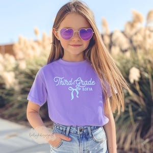 May include: A young person wearing heart-shaped sunglasses and a lavender t-shirt. The shirt has the words "Third Grade SOFIA" printed on it in a light blue script font with a bow graphic. The person is wearing blue jeans.